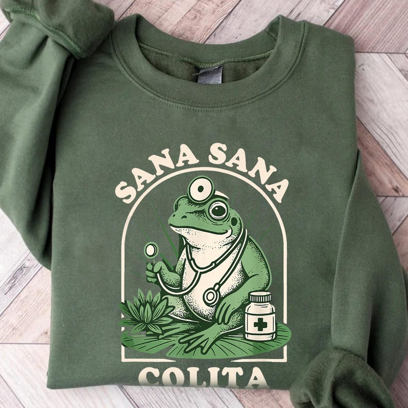 May include: Green sweatshirt featuring a cartoon frog doctor with a stethoscope and a pill bottle. The frog is inside a white arched frame with the words "Sana Sana Colita" in white text. The sweatshirt is a solid green color.