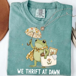 May include: A sage green t-shirt featuring a graphic of a frog holding a floral umbrella and a tote bag containing a framed picture and a book. The frog wears a yellow floral scarf. The text WE THRIFT AT DAWN is printed in white at the bottom.