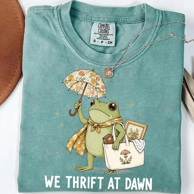May include: A sage green t-shirt featuring a graphic of a frog holding a floral umbrella and a tote bag containing a framed picture and a book. The frog wears a yellow floral scarf. The text WE THRIFT AT DAWN is printed in white at the bottom.