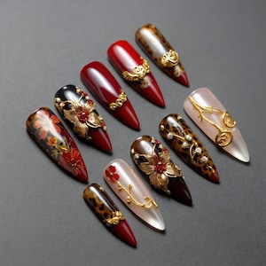 May include: A collection of ten long, almond-shaped artificial nails. The nails feature various designs, including solid colors, floral embellishments, leopard print, and gold accents. Some nails have 3D floral designs and rhinestones.