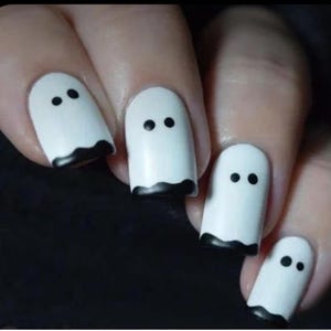 May include: White nail polish with black ghost designs. Each nail features a white base with two black dots for eyes and a black wavy line at the bottom, creating a spooky Halloween theme.