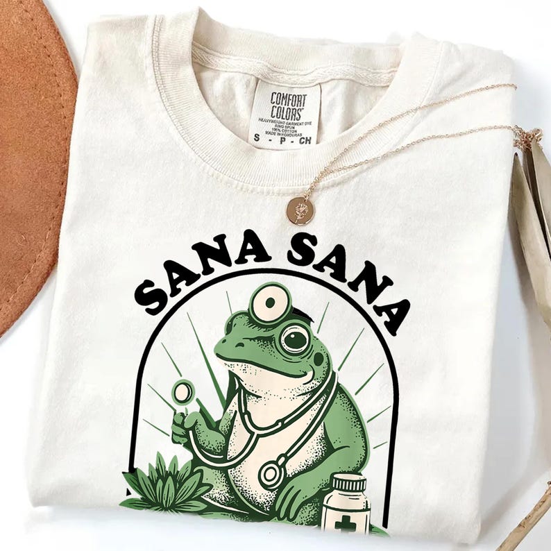 May include: Cream-colored t-shirt featuring a cartoon frog doctor with a stethoscope and the words "SANA SANA" in black. A gold necklace rests on the shirt. The frog is green and the design includes a medicine bottle.