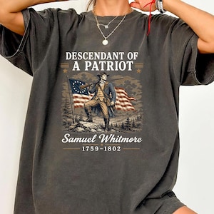 May include: Dark gray t-shirt featuring a patriotic design. The graphic includes an illustration of a man with a rifle standing in front of a flag. Text reads "DESCENDANT OF A PATRIOT" and "Samuel Whitmore 1759-1802".