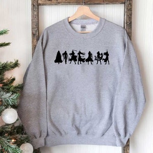 May include: A gray sweatshirt with a black silhouette of a group of women in various poses. The women are wearing different outfits and accessories, including capes, swords, and hats.