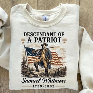 May include: Cream-colored sweatshirt featuring a graphic of a man in colonial attire holding a rifle, with an American flag in the background. Text reads "Descendant of a Patriot" and "Samuel Whitmore 1759-1802".