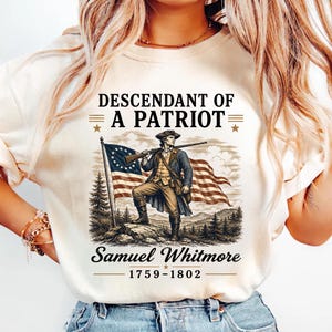 May include: Cream-colored t-shirt featuring a graphic of a colonial-era figure holding a rifle, standing before a waving flag. Text reads "DESCENDANT OF A PATRIOT" above and "Samuel Whitmore 1759-1802" below the image.