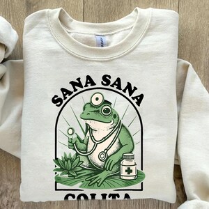 May include: Cream-colored sweatshirt with a graphic of a green frog doctor wearing a stethoscope and holding a medical tool. The text "SANA SANA COLITA DE RANA" is arched above and below the frog illustration.