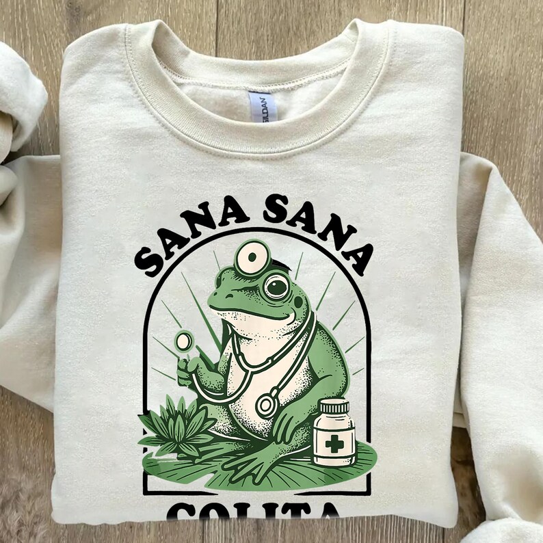 May include: Cream-colored sweatshirt with a graphic of a green frog doctor wearing a stethoscope and holding a medical tool. The text "SANA SANA COLITA DE RANA" is arched above and below the frog illustration.