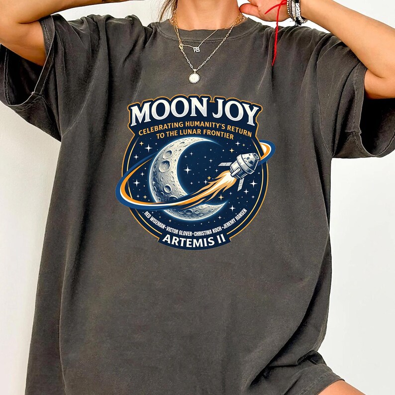 May include: Dark gray t-shirt featuring a circular graphic with the text "MOON JOY" and "CELEBRATING HUMANITY'S RETURN TO THE LUNAR FRONTIER." The graphic includes a crescent moon, a rocket, and the text "ARTEMIS II."