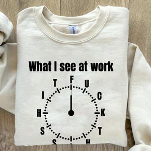 May include: Cream-colored sweatshirt with the text "What I see at work" in black. Below the text is a clock design with the letters T, F, U, C, K, T, H, I, S, and H. The sweatshirt is folded on a wooden surface.