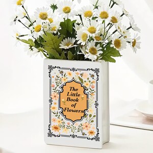 Può includere: Un vaso di ceramica bianco a forma di libro, con il titolo "The Little Book of Flowers". È pieno di fiori bianchi simili a margherite con centri gialli e foglie verdi. Il design del libro presenta elementi floreali e di api.