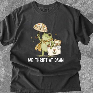 May include: A charcoal gray t-shirt featuring a graphic of a green frog holding a floral umbrella and a white tote bag with a mushroom print. The frog wears a yellow floral scarf and carries a framed picture. The text reads WE THRIFT AT DAWN.