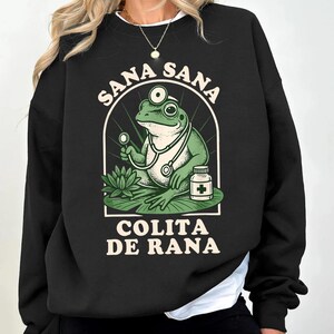 May include: Black sweatshirt featuring a green frog doctor with a stethoscope, holding a medical instrument, and a medicine bottle. The text "SANA SANA" arches above the frog, and "COLITA DE RANA" is below. The frog is on a lily pad.
