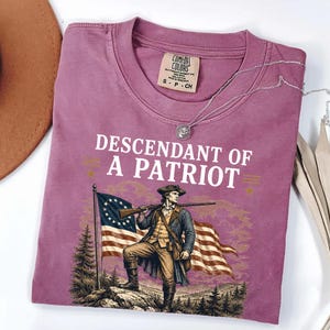 May include: A mauve Comfort Colors t-shirt with the text "DESCENDANT OF A PATRIOT" and an illustration of a colonial soldier holding a rifle in front of an American flag. A silver necklace is draped across the shirt.