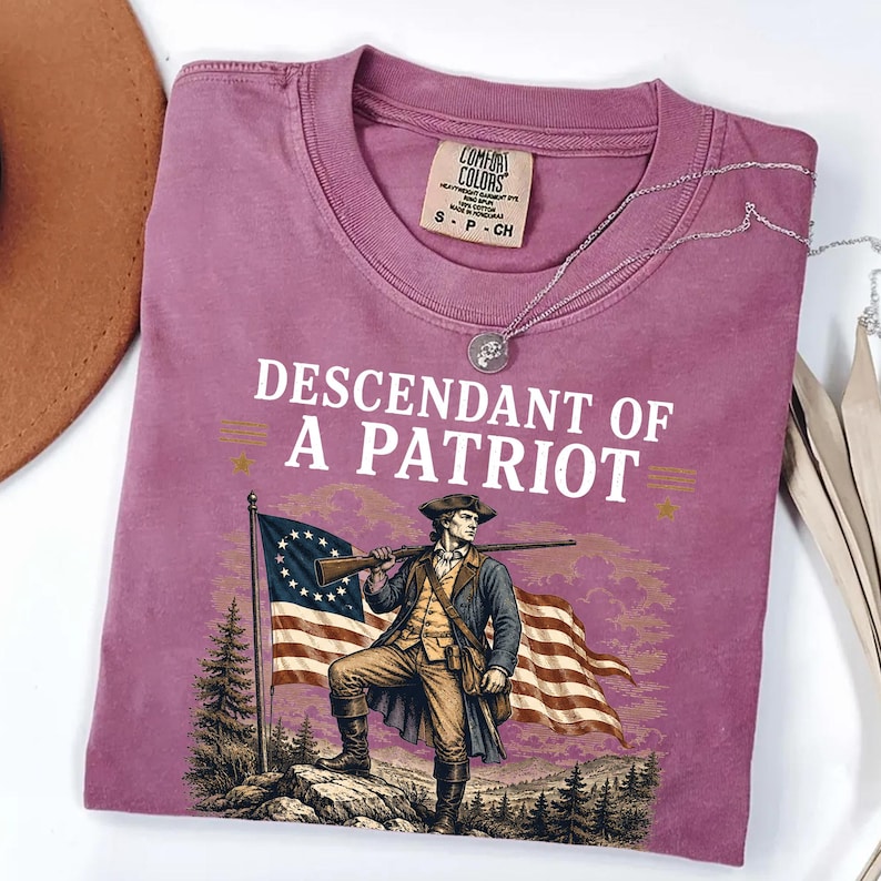 May include: A mauve Comfort Colors t-shirt with the text "DESCENDANT OF A PATRIOT" and an illustration of a colonial soldier holding a rifle in front of an American flag. A silver necklace is draped across the shirt.