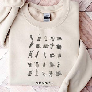 May include: A beige sweatshirt with a black and white graphic of various items used in the game Phasmophobia. The text "Phasmophobia" is printed in black at the bottom of the graphic.