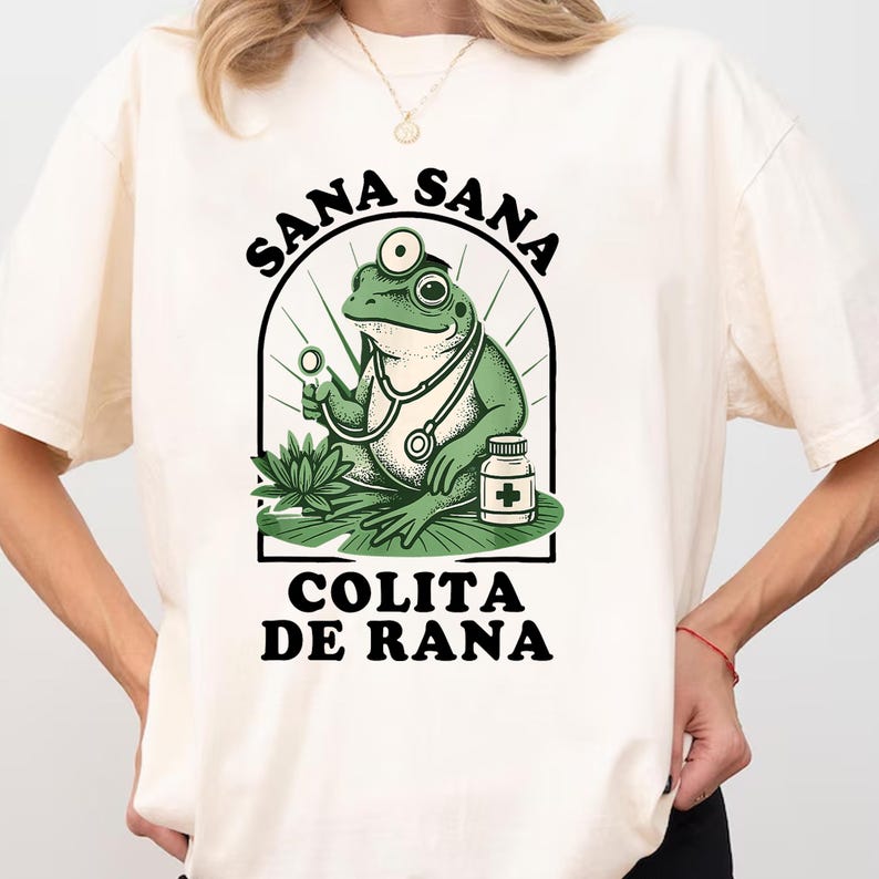 May include: Cream-colored t-shirt featuring a green frog doctor with a stethoscope, holding a medical instrument, and a medicine bottle. The text "SANA SANA COLITA DE RANA" is arched above and below the frog illustration.