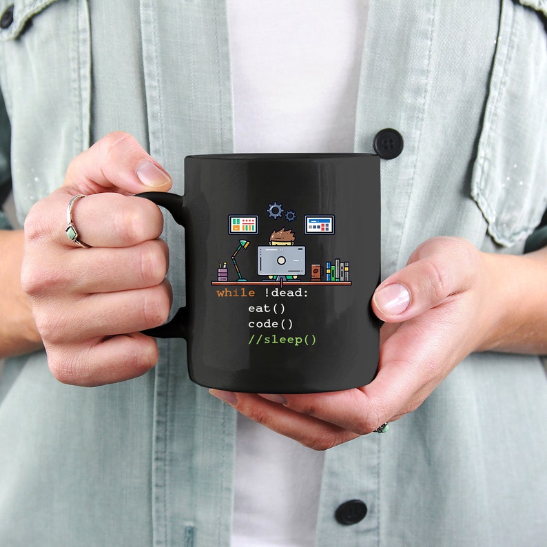 K&ouml;nnte beinhalten: Schwarze Kaffeetasse mit einem Design zum Thema Programmierung. Die Tasse hat einen Henkel und zeigt den Text "while !dead: eat() code() //sleep()" zusammen mit einer Grafik eines Programmierers und Computerbildschirmen.