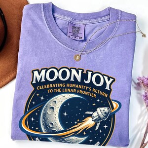 May include: A lilac-colored t-shirt with the words "MOON JOY" and a graphic of the moon, stars, and a spaceship. The shirt also has the text "CELEBRATING HUMANITY'S RETURN TO THE LUNAR FRONTIER". A silver necklace with a pendant is on top of the shirt.