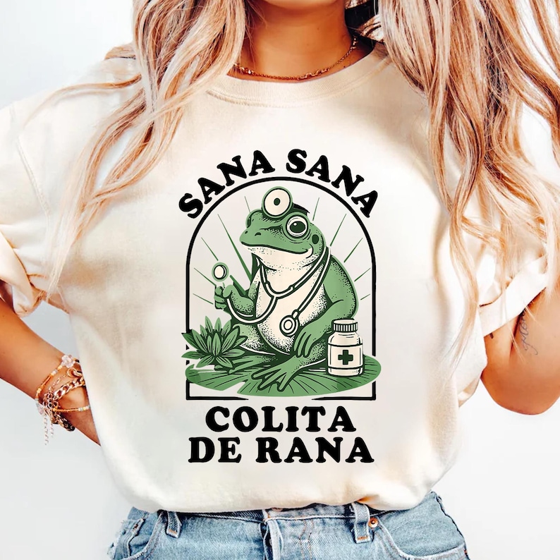 May include: Cream-colored t-shirt featuring a cartoon frog doctor with a stethoscope, holding a medical tool. The text "SANA SANA" arches above the frog, with "COLITA DE RANA" below. The frog is green, and the text is black.