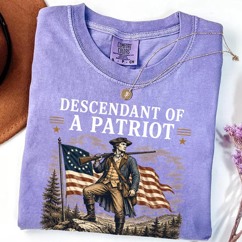 May include: A lilac-colored t-shirt with the text "DESCENDANT OF A PATRIOT" and an illustration of a colonial soldier holding a rifle in front of an American flag. A gold necklace with a pendant is draped across the shirt.