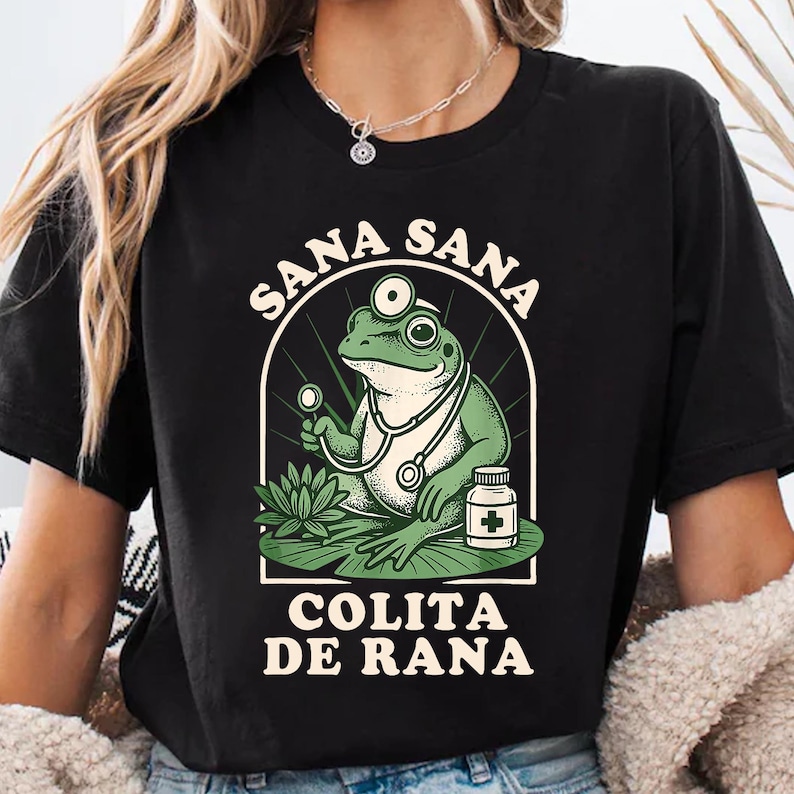May include: Black t-shirt featuring a cartoon frog doctor with a stethoscope, holding a medical instrument, and a medicine bottle. The text "SANA SANA COLITA DE RANA" is arched above and below the frog illustration.