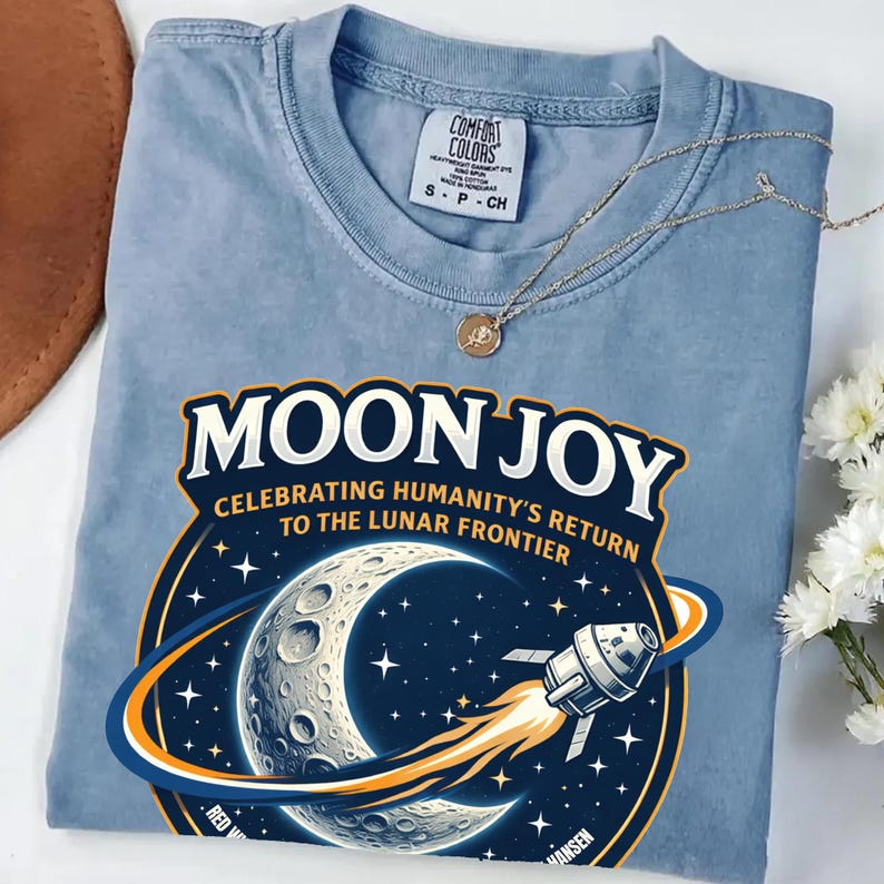 May include: A light blue t-shirt with the words "MOON JOY" in a bold font, celebrating humanity's return to the lunar frontier. The graphic includes a detailed illustration of the moon, stars, and a spaceship. A gold necklace with a pendant rests on the shirt.