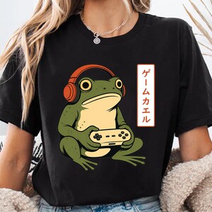 Puede incluir: Una camiseta negra de manga corta con un gráfico de una rana verde que lleva auriculares naranjas y sostiene un mando de juego. A la derecha de la rana hay un recuadro blanco vertical que contiene el texto japonés ゲー ムカエル en color rojo.