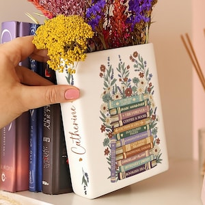May include: A white ceramic vase shaped like a book, decorated with floral and book-themed illustrations. The vase is filled with colorful dried flowers. The name "Catherine" is written on the side.