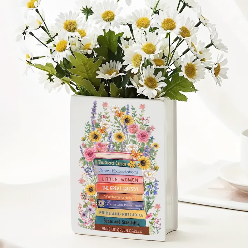 May include: White ceramic vase shaped like a stack of books, with titles including "The Secret Garden" and "Pride and Prejudice." The vase is decorated with colorful flowers and holds a bouquet of white daisies with yellow centers.