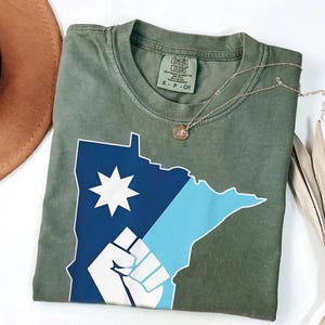 May include: An olive green t-shirt with a graphic of the state of Minnesota in blue and white, including a star and a fist design. A silver necklace with a small pendant rests on the shirt. The shirt is labelled "Comfort Colors".