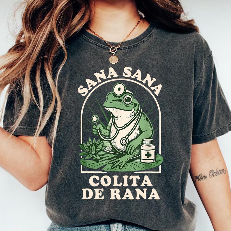 May include: Dark gray t-shirt featuring a cartoon frog doctor with a stethoscope, holding a medical instrument, and a pill bottle. The text "SANA SANA COLITA DE RANA" is printed above and below the frog illustration.