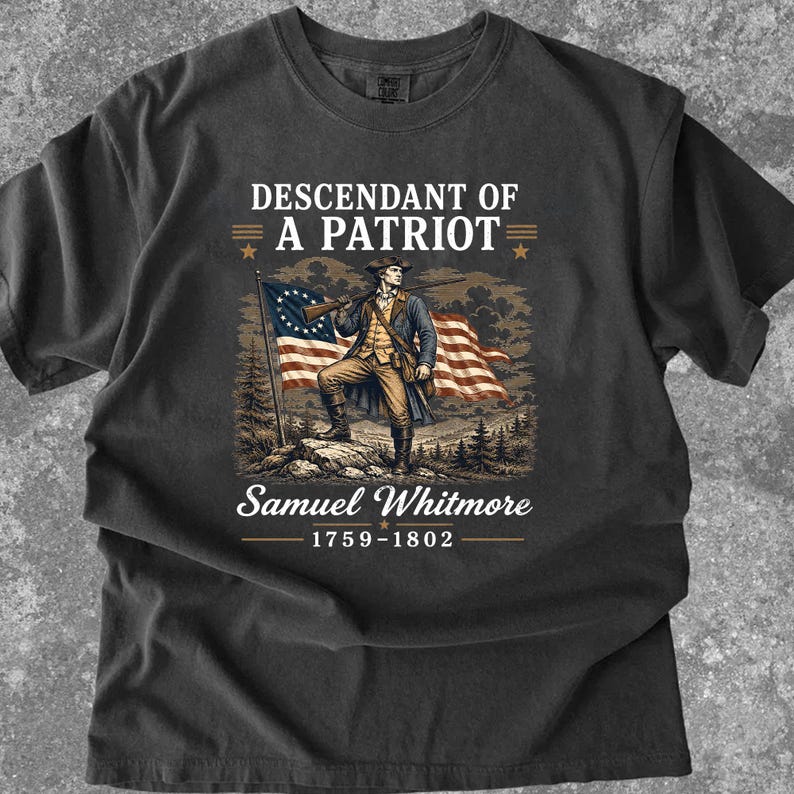 May include: Dark gray t-shirt featuring a patriotic design. The graphic includes a colonial-era figure with a rifle, the American flag, and the text "Descendant of a Patriot" and "Samuel Whitmore 1759-1802".
