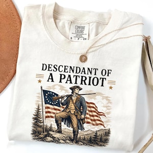 May include: Cream-colored t-shirt with the text "DESCENDANT OF A PATRIOT" above an illustration of a colonial soldier holding a rifle in front of an American flag. A gold necklace with a round pendant is draped on the shirt.