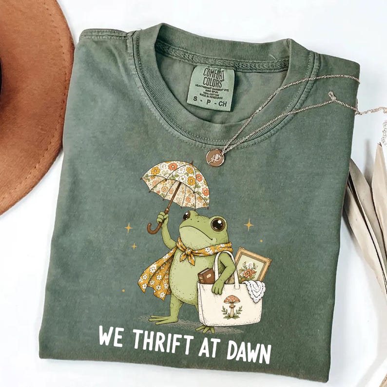May include: An olive green t-shirt featuring a graphic of a frog holding a floral umbrella and carrying a tote bag with a mushroom print. The text "WE THRIFT AT DAWN" is printed below the frog. The shirt has a Comfort Colors brand label at the neck.