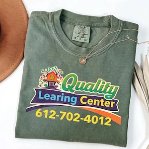 May include: A sage green Comfort Colors t-shirt with the words "Quality Learning Center" in a colorful graphic design. The phone number 612-702-4012 is printed below the logo. A silver necklace with a small pendant rests on the shirt.