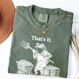 May include: Green t-shirt with the text "That's it." and a cartoon frog holding a pitchfork. The frog is wearing an apron and standing in front of a wheelbarrow and a crate filled with produce. A gold necklace and a brown hat are also visible.