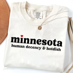 May include: Cream-colored t-shirt with the word "minnesota" in black, a red heart, and the text "human decency & hotdish." A gold necklace with a pendant rests on the shirt. The shirt has a tag that says "Comfort Colors."