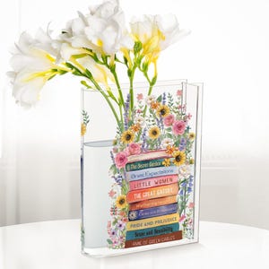 May include: A clear rectangular vase with a floral design and a stack of book titles. The vase holds white and yellow flowers. The book titles include "The Secret Garden," "Great Expectations," and "Little Women."