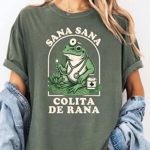 May include: Green t-shirt featuring a cartoon frog doctor with a stethoscope, holding a medical instrument, and a medicine bottle. The text "SANA SANA COLITA DE RANA" is arched above and below the frog illustration.
