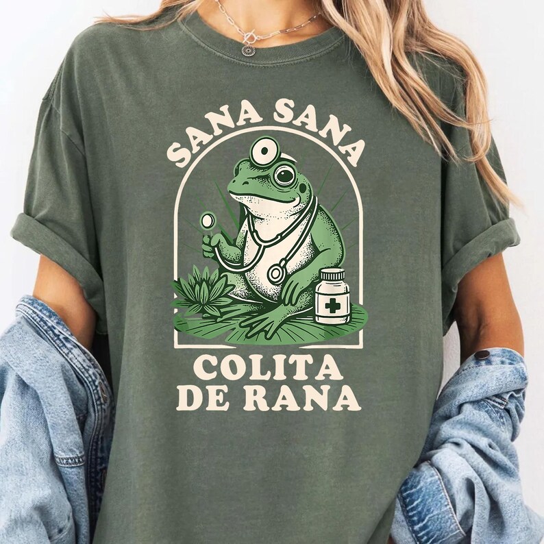May include: Green t-shirt featuring a cartoon frog doctor with a stethoscope, holding a medical instrument, and a medicine bottle. The text "SANA SANA COLITA DE RANA" is arched above and below the frog illustration.