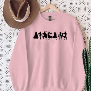May include: A pink sweatshirt with a black silhouette of a group of women wearing capes and holding swords. The text "AMAZON" is printed below the silhouettes.