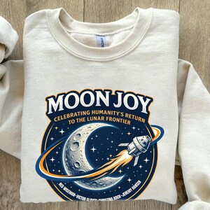 May include: Cream-colored sweatshirt with a graphic featuring the words "MOON JOY" above a crescent moon, stars, and a rocket ship. The graphic celebrates humanity's return to the lunar frontier.