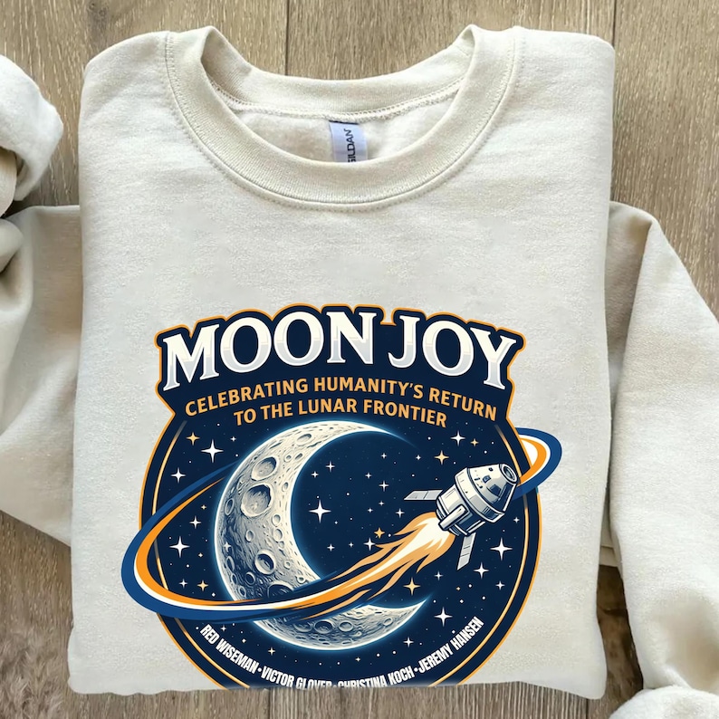 May include: Cream-colored sweatshirt with a graphic featuring the words "MOON JOY" above a crescent moon, stars, and a rocket ship. The graphic celebrates humanity's return to the lunar frontier.
