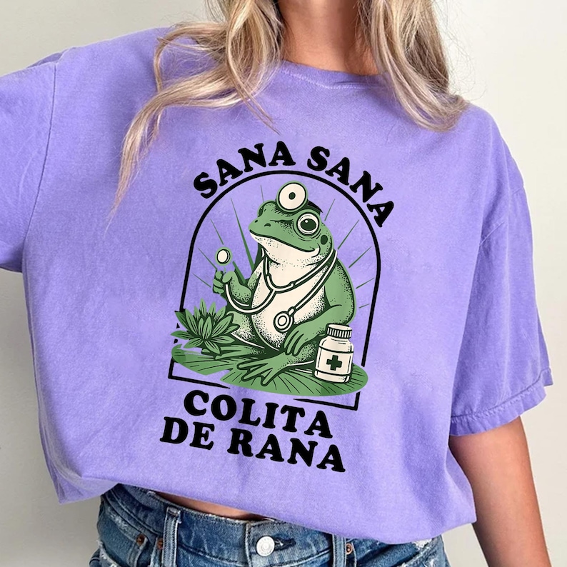 May include: A lilac-colored t-shirt featuring a cartoon frog doctor with a stethoscope, glasses, and a pill bottle. The frog is inside a black arched frame with the words "Sana Sana" above and "Colita de Rana" below.