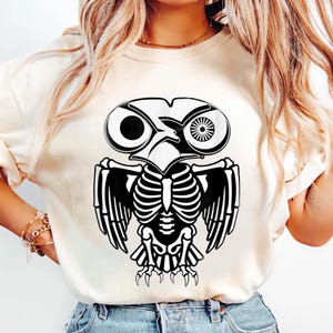May include: Cream-colored t-shirt featuring a black and white owl skeleton design. The graphic includes the owl's head, wings, and skeletal structure. The design is centered on the shirt, creating a striking visual.
