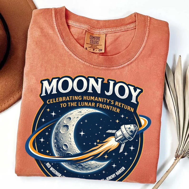 May include: A coral-colored t-shirt featuring a graphic with the words "MOON JOY" above an illustration of the moon, stars, and a spaceship. The text "CELEBRATING HUMANITY'S RETURN TO THE LUNAR FRONTIER" is also included.