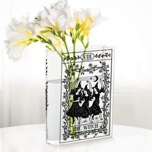 May include: A clear glass vase with a black and white tarot card design featuring three witches dancing under a crescent moon. The vase holds white and yellow flowers.
