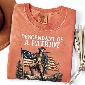 May include: Coral-colored Comfort Colors t-shirt with the text "DESCENDANT OF A PATRIOT" above an illustration of a colonial soldier holding a rifle and standing in front of a waving American flag.