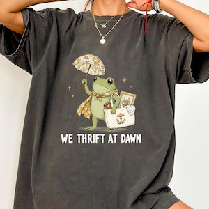 May include: A charcoal gray t-shirt featuring a graphic of a green frog holding a floral umbrella and a white tote bag with a mushroom illustration. The frog wears a floral scarf. Below the graphic, the text reads WE THRIFT AT DAWN in white.